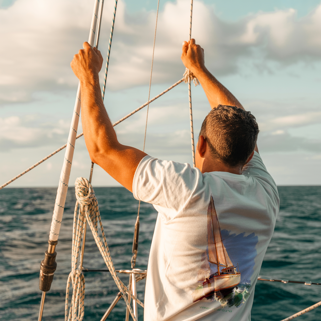 A Wear-it-around Anchored in Freedom collection image showing a man on sailboat in a white t-shirt that features a painting of sailboat in high seas.